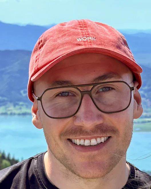 Florian Erwerth lacht in die Kamera. Er trägt eine Brille und eine rote Kappe, hinter ihm liegen ein See und Berge.